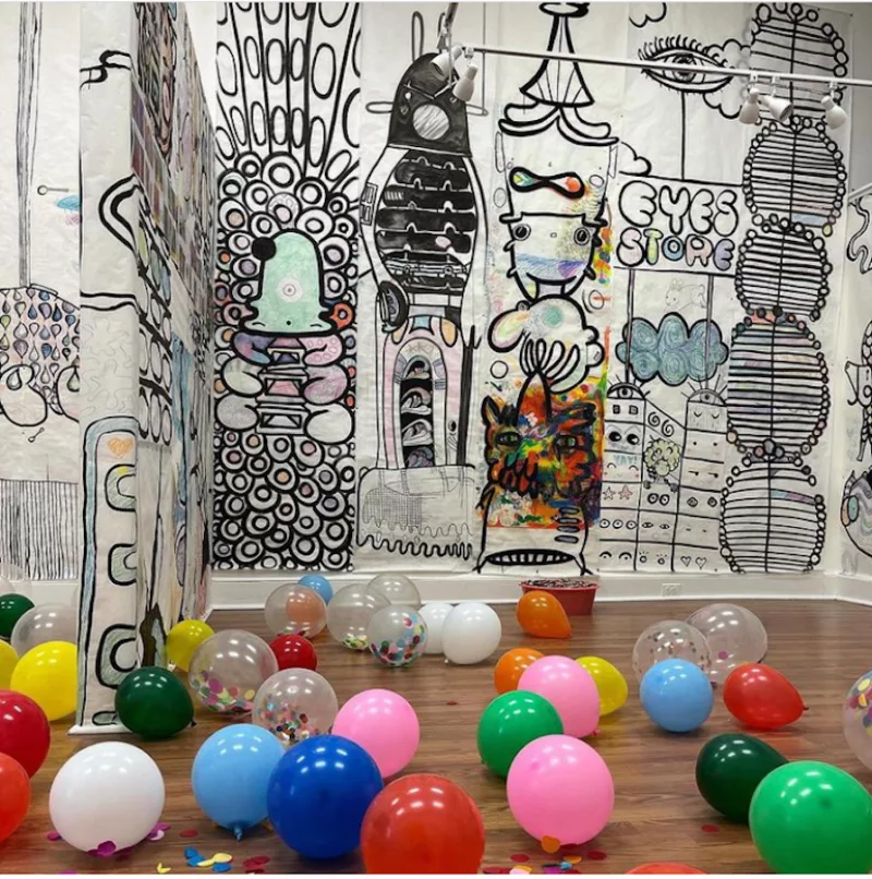 A gallery with two walls covered with coloring book-like imagery that has been colored in and a floor covered with balloons of many colors